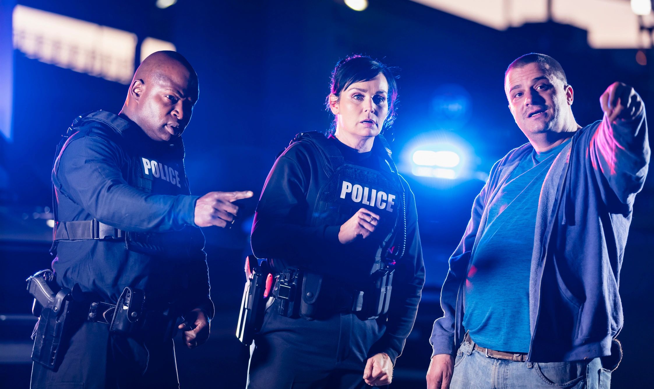 Two police officers speaking with a man at night near a patrol car with emergency lights.